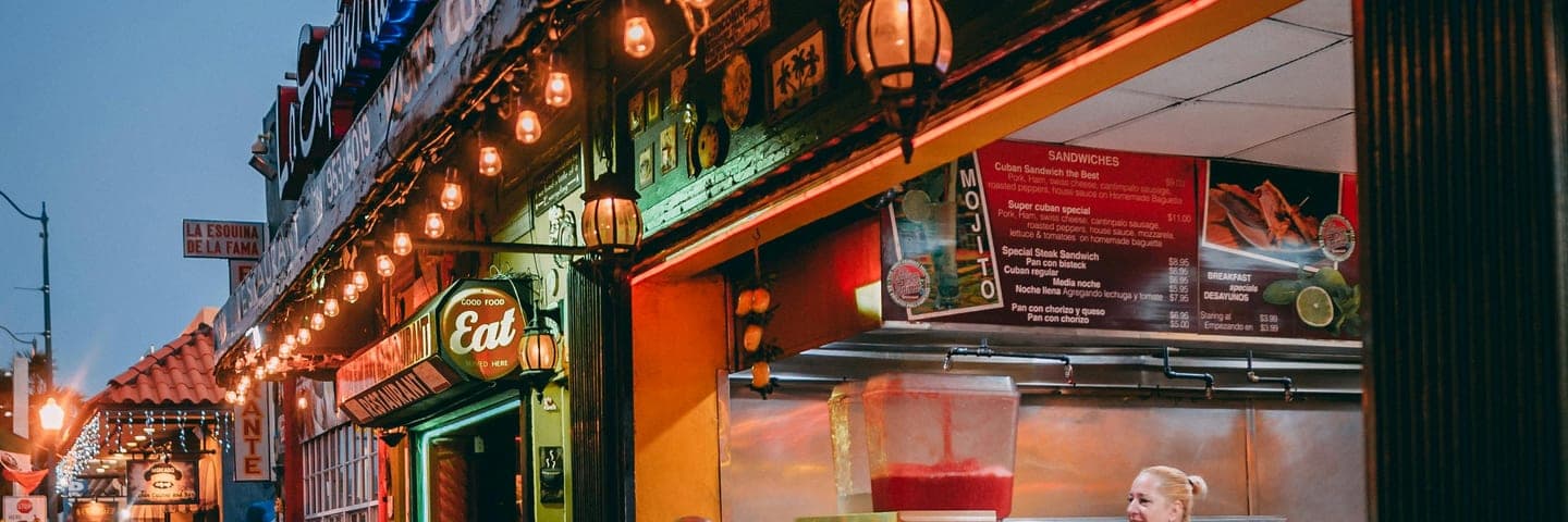 La Esquina de la Fama on Calle Ocho — a Cuban restaurant in Miami's Little Havana, warm string lights glowing at dusk, bilingual menus, a worker at the counter